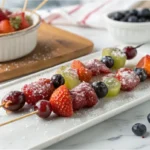 A close-up of glossy Tanghulu skewers with strawberries, grapes, and blueberries, coated in hardened sugar, placed on a white marble countertop in a bright white kitchen.